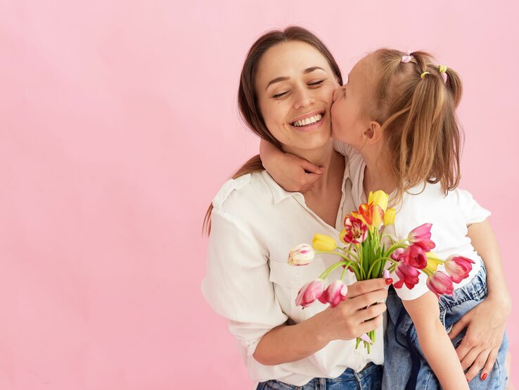 Junge Mutter mit braunem Haar in weißer Bluse lacht, während ihr Kind sie küsst und Tulpen hält, vor rosa Hintergrund.